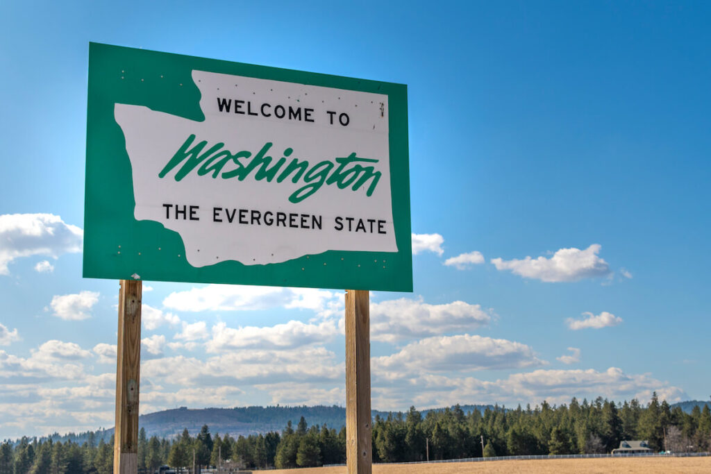 A roadside sign with welcome to Washington State, the evergreen state written on it with a rural setting and blue sky behind.