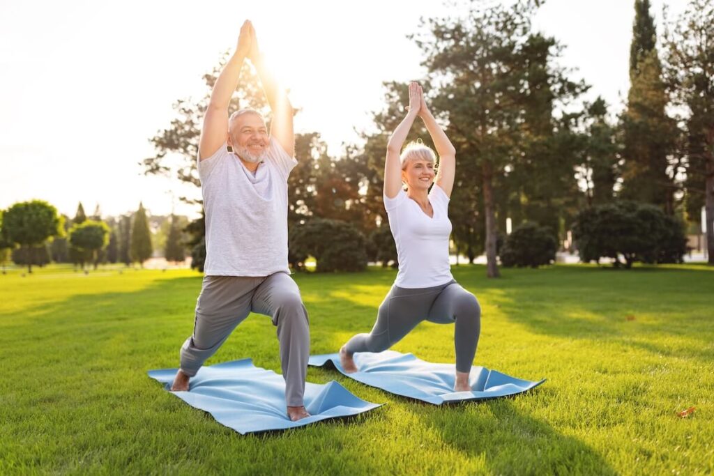 Happy positive fit mature couple of senior woman and man practicing partner yoga on open fresh air outside standing barefoot