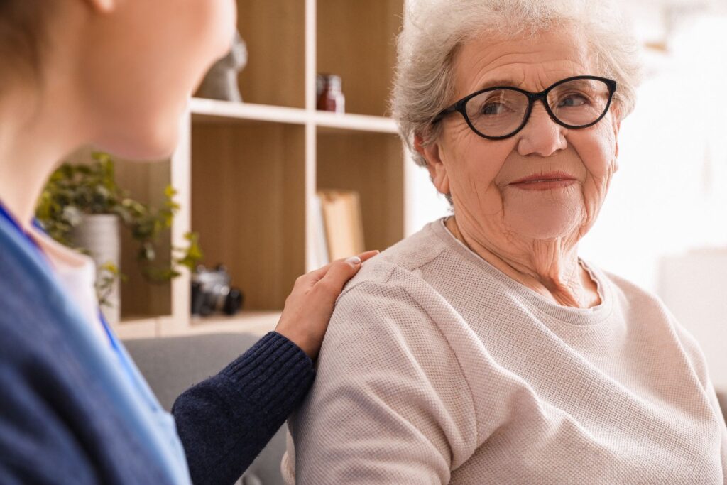 Knowing When to Ask for Help as an older woman sits on a couch while a supportive caregiver gently rests a hand on her shoulder during a calm conversation.