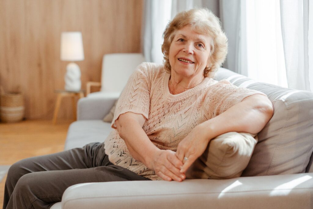 Independent living as an older woman relaxes comfortably on a sofa in a bright, well-kept living space, smiling with ease and confidence.
