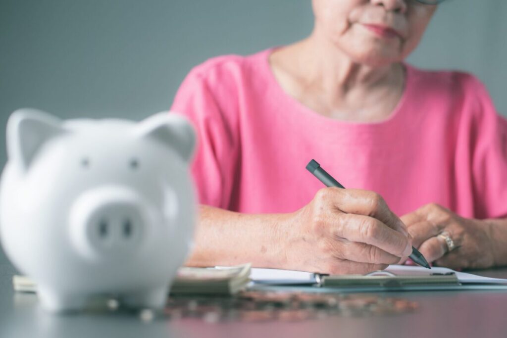 Senior woman reviewing finances beside a piggy bank illustrating the hidden cost of aging in place rather that at Village Green