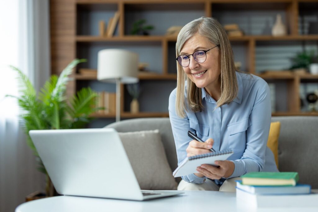 Older woman smiling while taking notes on a laptop at home, representing lifelong learning.