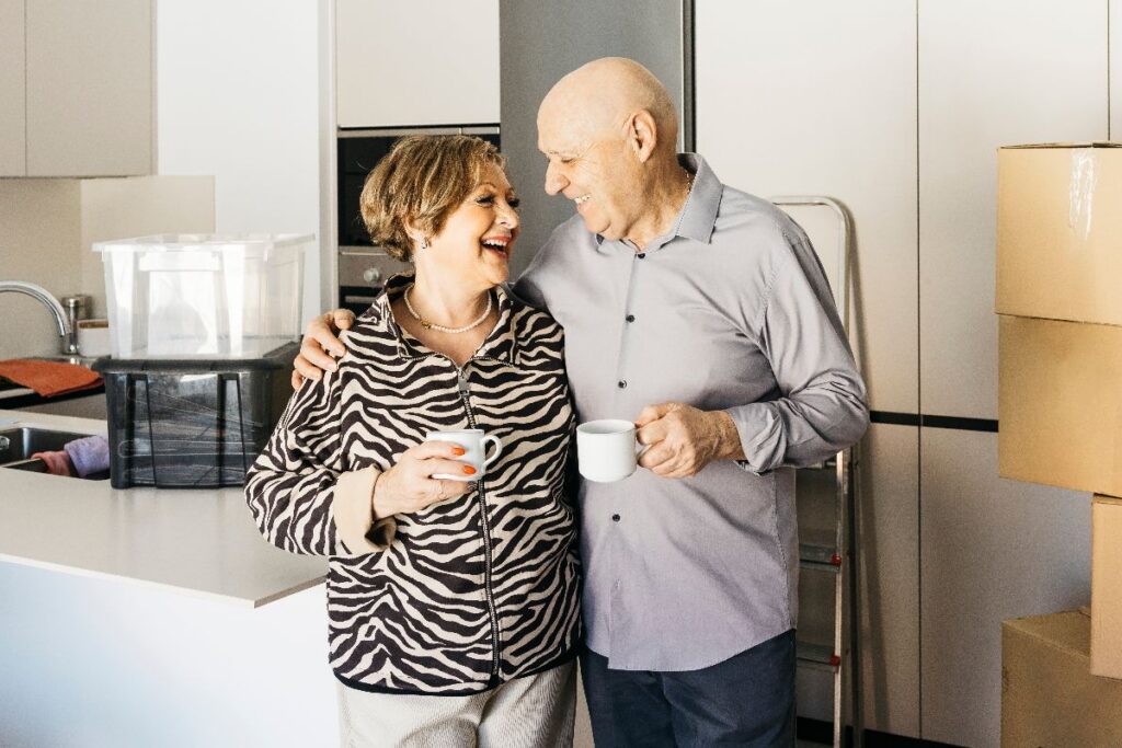 Happy older couple standing in their kitchen with moving boxes, symbolizing downsizing tips for seniors.