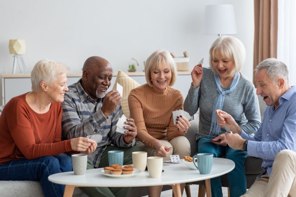 Diverse group of seniors laughing and playing cards together, enjoying community at Village Green Senior Living retirement community