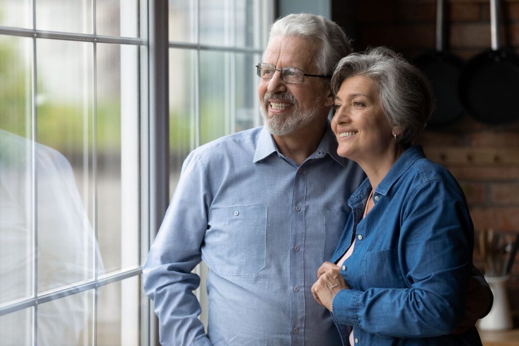 An older couple stands close together by a large window, smiling warmly as they look out at the view, the man's arm around the woman as she holds onto his — a peaceful, connected moment that reflects the vibrant yet serene lifestyle of independent living in West Seattle.