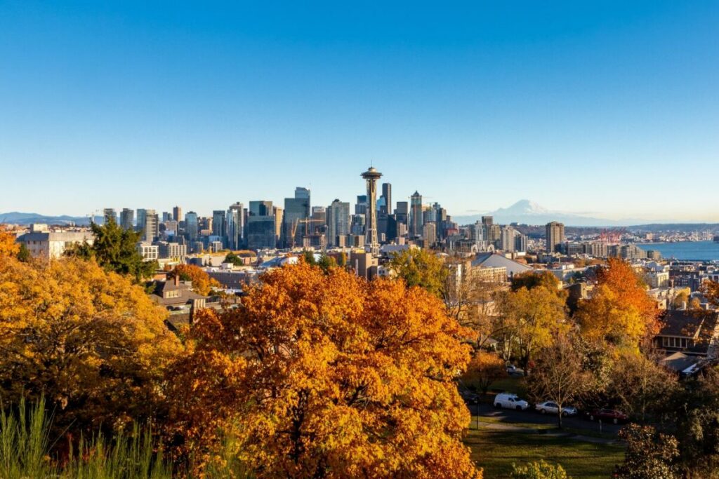 A panoramic view of the Seattle skyline and Space Needle rises beyond vibrant orange and gold autumn foliage, with Mount Rainier visible in the distance across the water — a scenic backdrop that captures the natural beauty surrounding Pacific Northwest senior living options this time of year.