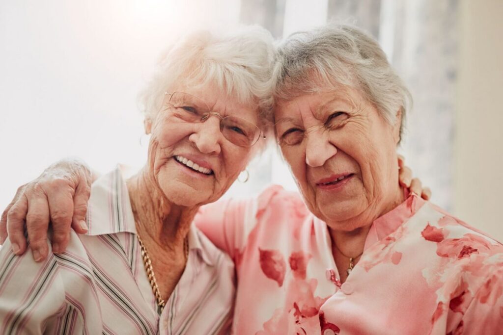 Two elderly women sitting closely together, smiling warmly, symbolizing companionship and addressing themes of loneliness in older adults.