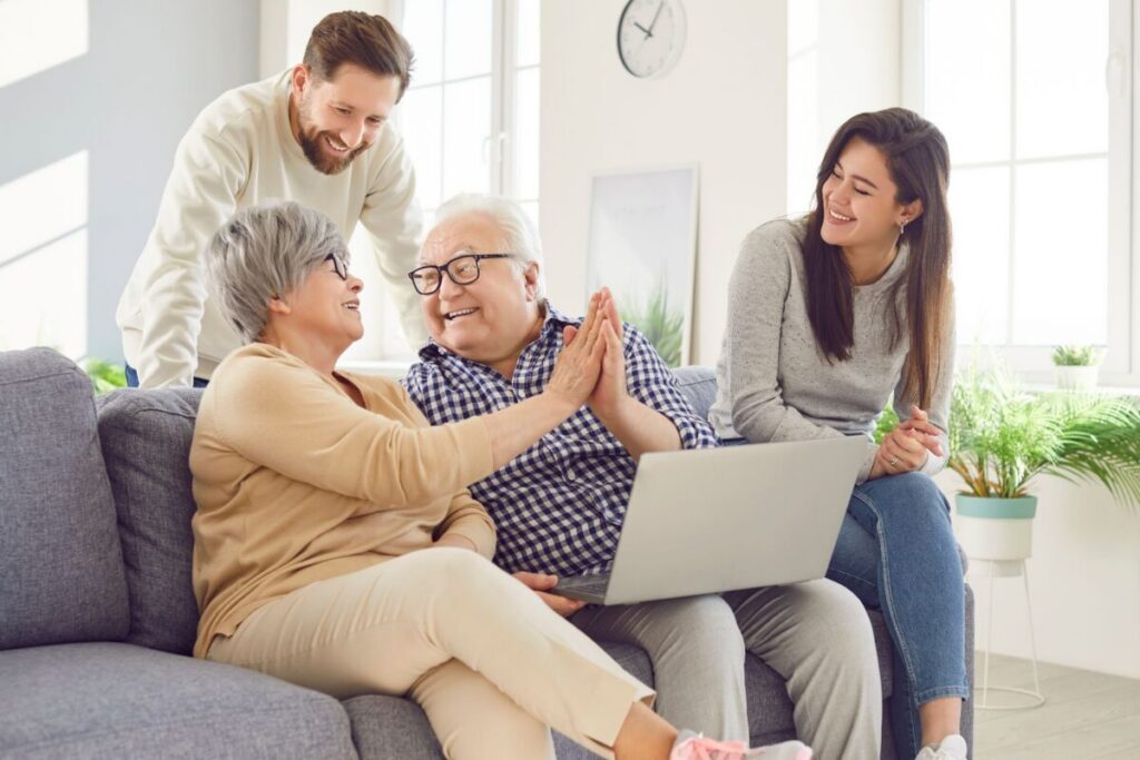 A joyful multi-generational family moment in a modern living room, featuring an elderly couple on a gray sofa celebrating with a high-five while using a laptop, accompanied by two younger family members smiling warmly. Perfectly illustrating themes of family bonding and support, ideal for "assisted living near me" services.